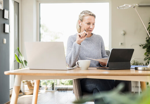 Businesswoman with digital tablet and laptop holding paper while working at home office