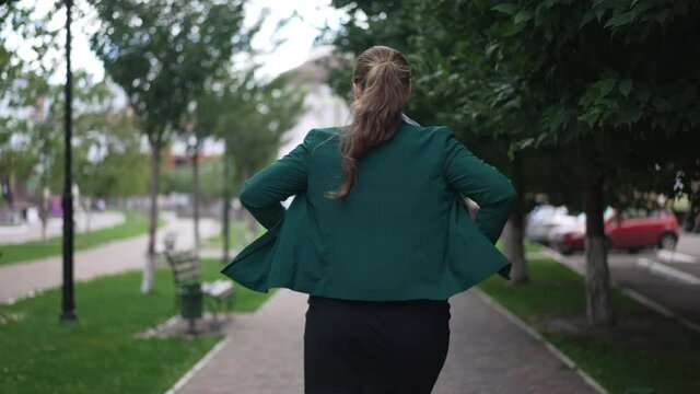 Back view obese woman walking away in slow motion along city sidewalk. Confident carefree Caucasian overweight plus-size lady strolling on street outdoors looking around