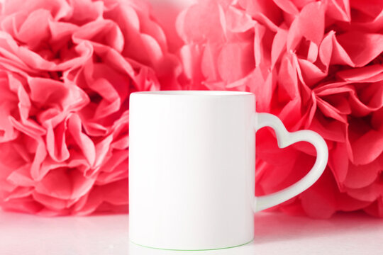 White mug on red paper flowers background with heart shaped handle for Valentine's day or Mothers day, template for your design.