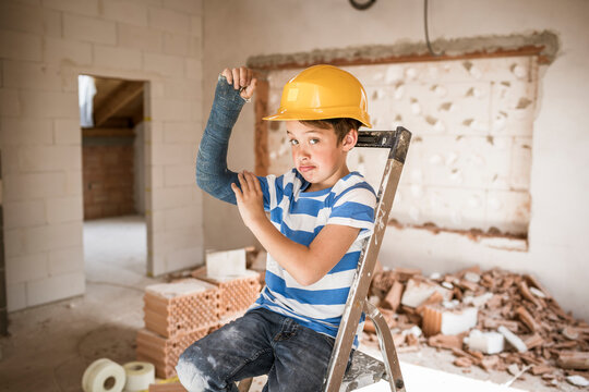 Boy flexing broken arm muscles while sitting on ladder during house renovation