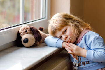 Upset toddler girl sitting by window and looking out. Sad crying child feeling unhappy. Lockdown...