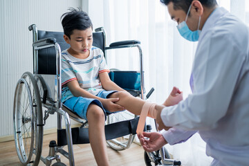 Obraz premium Asian boy sitting on a wheelchair is having the doctor wrap a cloth around his leg because he broke his leg at the hospital.
