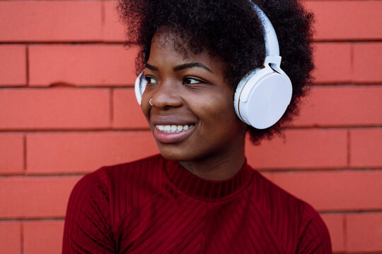 Smiling Afro woman with headphones looking away by brick wall