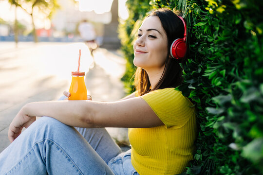 Smiling woman with juice bottle listening music through headphones while leaning on hedge