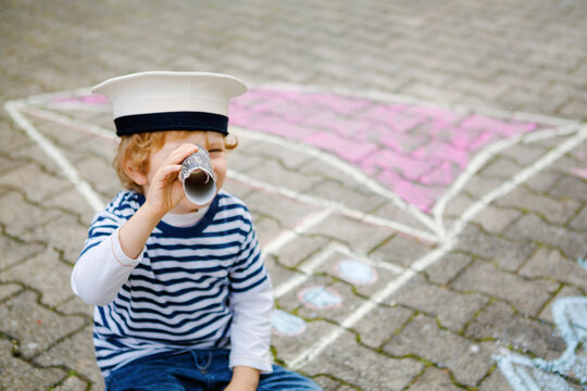 Little kid boy as pirate on ship or sailingboat picture painting with colorful chalks on asphalt. Creative leisure for children outdoors in summer. Child with captain hat and binoculars.