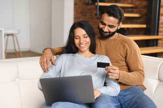 Cheerful Indian couple watching at laptop screen and checking credit card number to make online transaction. Smiling spouses having shopping online. Transferring money, online banking concept