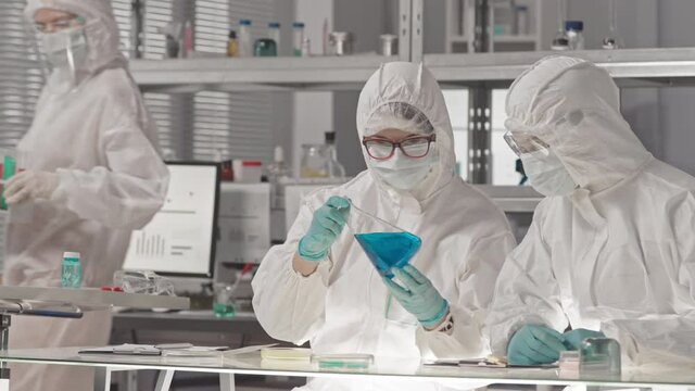 Waist-up of male Biracial and female Caucasian laboratory workers wearing hazmat overalls, goggles and masks, sitting in research center, analyzing hazardous chemicals in test tubes