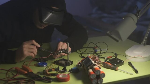 man in black sweatshirt in dark room is soldering and assembling a bomb of dynamite with telephone as detonator