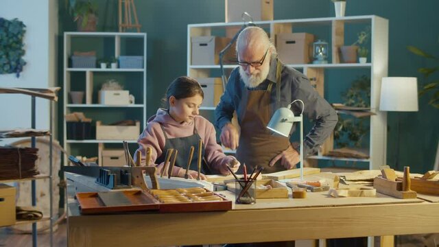 Grandfather Carpenter with his Teenage Granddaughter Works in the Workshop, Making Artistic Woodcarving. Communication of Generations. Handicraft, Skills Training. Profession, Art and Hobby Concept.