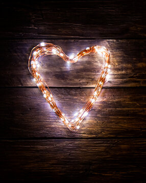 A heart shaped figure laid out light LED garland on vintage brown wooden board. Heart made of LED rice lights. Concept of Valentine Day, strong eternal love, light of love in the heart of a lover