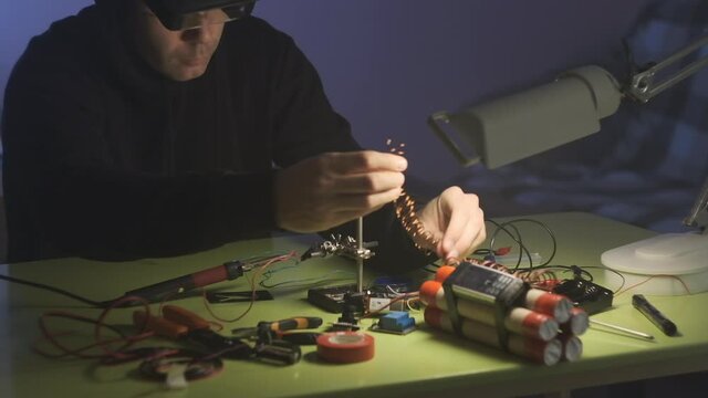 man in black sweatshirt in dark room is soldering and assembling a bomb of dynamite with telephone as detonator
