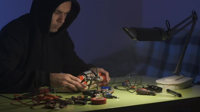man in black sweatshirt in dark room is soldering and assembling a bomb of dynamite with telephone as detonator