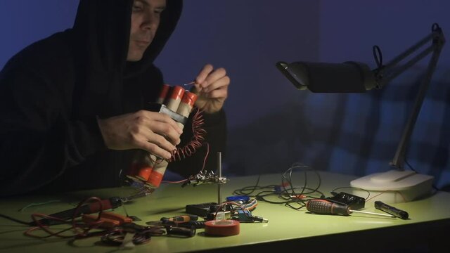 man in black sweatshirt in dark room is soldering and assembling a bomb of dynamite with telephone as detonator