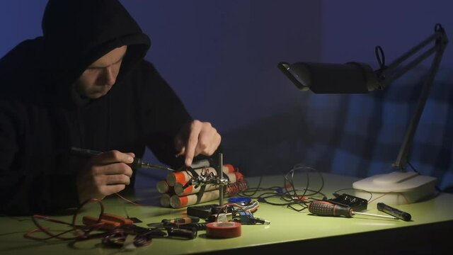 man in black sweatshirt in dark room is soldering and assembling a bomb of dynamite with telephone as detonator