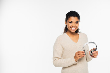 Fototapeta premium Smiling african american woman in sweater holding headphones isolated on white.