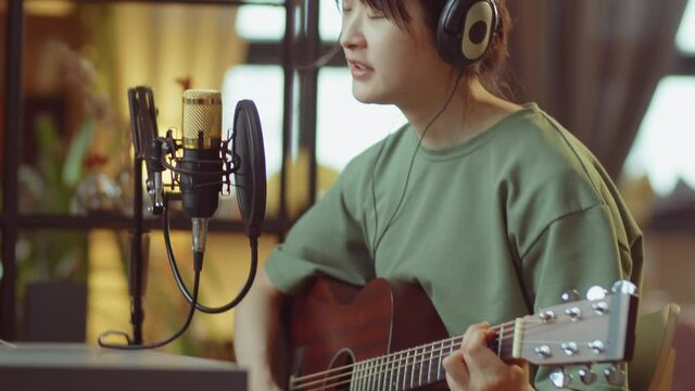Young Asian woman in headphones playing guitar and singing in microphone at home recording studio