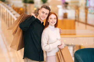 Portrait of happy beautiful young couple holding shopping paper bags with purchases and looking at camera, standing embracing in mall with festive bright interior, blurred background.