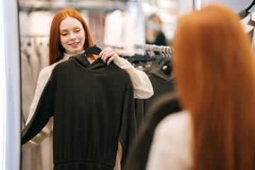 Portrait of smiling young woman buyer choosing clothes and looking to mirror in clothing store. Happy shopaholic female standing in front of mirror, holding dress and looking at herself.
