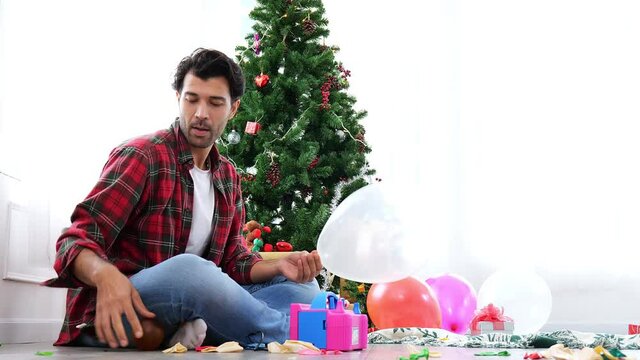 A handsome young Caucasian father blows balloons to decorate a room for Christmas party and has a naughty toddler nearby.There was a Christmas tree with twinkling lights in the corner of the room.