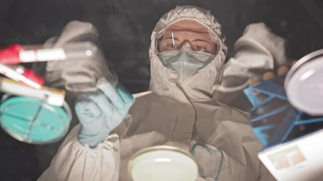 Low angle view through glass table top of female Caucasian scientist wearing hazmat suit, gloves, goggles and medical mask, standing in dark research center, working with Petri dish