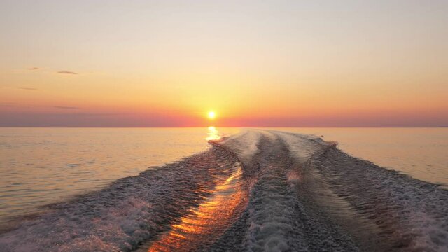 Fast boat rush on flat water of large lake, camera look back to vibrant sunset, orange sun hang low over horizon. Beautiful end of some journey or story. Speedboat make slight turns left and right