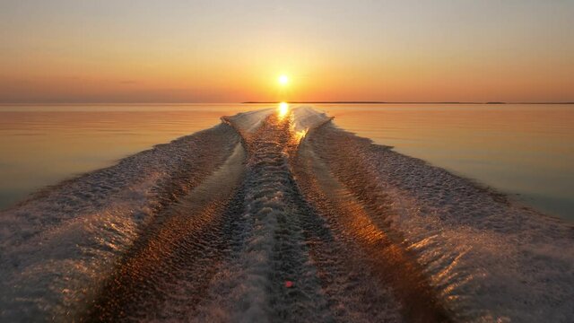 Look back to vivid sunset from speedboat running fast at calm water of large lake, cinematic shot. Wake and splashes behind motor boat, bright golden reflection of sun on water