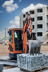 A mini excavator bucket rests on a pallet of paving slabs at a construction site. Compact construction equipment for earthworks and landscaping.