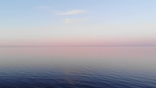 Pale colours of sky reflecting in water surface, scenic shot of Ladoga lake after sunset. Delicate pink and light blue shades blend from sky to horizon line. Small waves on water, aerial shot