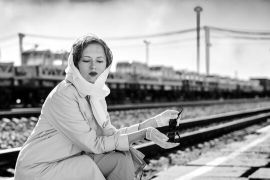 A young woman in a raincoat with sunglasses in her hands sits on the platform of a railway station. Retro style black and white outdoor shot