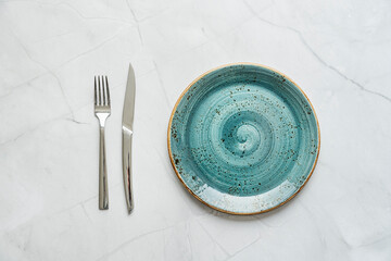Beautiful blue porcelain plate with stainless steel knife and fork on white marble surface - obrazy, fototapety, plakaty