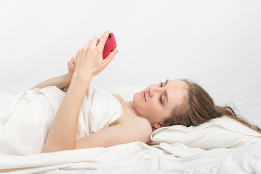 Young Woman Lies In Bed With Phone And Corresponds With Friends. Social Media Addiction. Side View. White Background