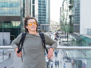 Young man with yellow eyeglasses and long hair stands on big city background. Modern buildings with glass wall. Millennial lifestyle.