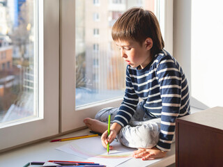 Little boy sits on window sill and draws rainbow with colored pencils. Creative leisure activity for children at home.