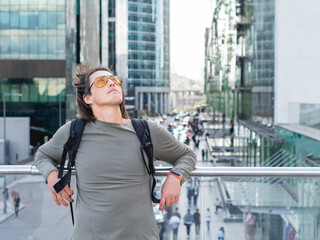 Young man with yellow eyeglasses and long hair stands on big city background. Modern buildings with glass wall. Millennial lifestyle.