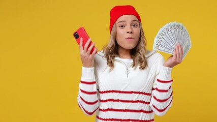 Excited joyful charming fun young girl teen student wears striped white shirt hat using mobile cell phone hold fan of cash money in dollar banknotes isolated on plain yellow background studio portrait
