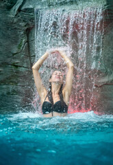 beautiful young sexy woman under the splashing water of the waterfall in the Spa Wellness pool, raises her arms, closes her eyes under the rippling water