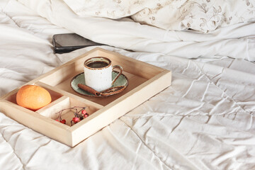 Morning coffee cup in bed, a mobile phone  on white blanket, cup of coffee, a tangerine, cinnamon stick and dried rowanberries. Autumn winter coffee time, copy space
