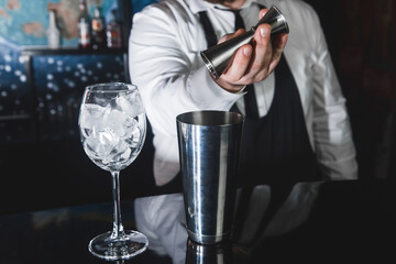 The hands of a professional bartender pour syrup into a measuring glass of jigger in a metal tool for preparing and stirring alcoholic cocktails of shaker drinks, and glasses with ice cubes
