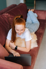 Vertical shot of happy young woman lying on comfortable sofa with white pretty Spitz pet dog and using laptop in living room. Happy redhead female spending time together with cute little doggy at home