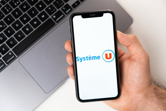 Systeme U mobile application on the smartphone screen. A mobile phone in a man hand near an open laptop. White background. November 2021, San Francisco, USA