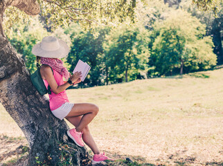 side view of a hiker dressed in pink and white, leaning against a tree while reading a travel diary. concept of life in nature and ecology.