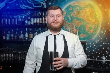 A bearded adult man of European appearance, a professional bartender, holds in his hands a tool for preparing and mixing alcoholic cocktails in a nightclub