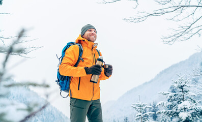 Smiling Man under snowfall dressed bright orange softshell jacket with a hot drink thermos flask...