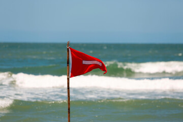 Red flag lifeguard danger warning sign on the beach. No swimming it's forbidden symbol, dangerous rip tide currents on water, enter at your on risk. Blue sunny sky and turquoise waves on background