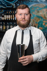 A bearded adult man of European appearance, a professional bartender, holds in his hands a tool for preparing and mixing alcoholic cocktails in a nightclub