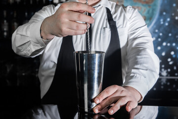 The hands of a professional bartender interfere with a bar spoon the contents of a metal shaker tool for preparing and stirring alcoholic cocktails