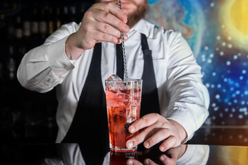 The hand of a professional bartender stirs the red syrup in an alcoholic cocktail with a bar spoon...