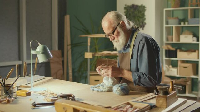 A Concentrated Elderly Man Makes Handicrafts, Knits a Product, Enjoys Leisure, Relaxes in a Cozy Workshop with a Warm Light. DIY Knitting, Handicraft, Creativity, Hobby Concept.