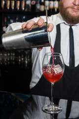 Professional bartender with a beard pours liquid into a glass of ice cubes from a tool for mixing and preparing alcoholic beverages cocktails metal shaker