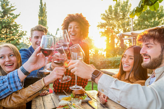 Group of multiethnic friends living healthy lifestyle and smiling and joking while drinking red wine at outdoor pub restaurant - Young people toasting with wine glass during happy hour at bar